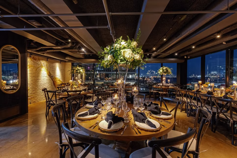 A tall floral centerpiece above a set round table, skyline through the windows at night